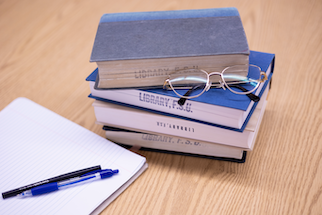 A stack of three books on a wooden surface, with the topmost book partially covered by a pair of glasses. Below the books, there is an open notebook with a blue pen resting on it. The titles of the books are not fully visible due to the angle and positioning of the glasses. This image represents materials commonly used in research, such as academic books for reference, a notebook for taking notes or organizing thoughts, and glasses that imply reading or detailed work.